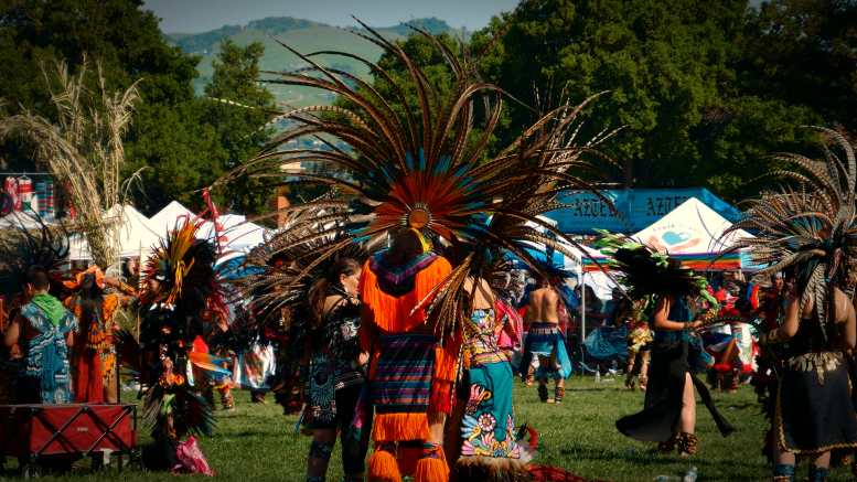 Mexica dancers in regalia perform together in a grass circle